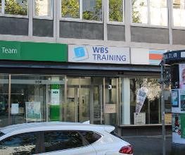 Glas-Eingang mit Schild der WBS TRAINING an einer Straße. Rechts eine Litfasssäule.