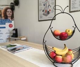 Gemütliche Küche/Aufenthaltsbereich. Heller Holztisch mit Zeitschriften und einer großen Obstschale mit frischem Obst. An den Wänden Bilder, im Hintergrund ein Aufsteller.