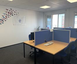Schulungsraum mit mehreren Arbeitsbereichen, die durch blaue Trennwände voneinander abgegrenzt sind und mit 2 PC-Bildschirmen, Tastatur, Maus und Headset ausgestattet sind. Fensterreihe und gebastelte Schmetterlinge an der Wand.