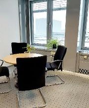 Raum mit einem Tisch und 4 schwarzen Stühlen. Links hängt ein Plakat an der Wand, rechts steht eine Zimmerpflanze auf dem Fensterbrett. Der Boden ist mit Teppich ausgelegt.