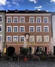Außenansicht eines Gebäudes mit brauner Fassade und der Aufschrift „Köllerbräu“. Davor befinden sich Sitzplätze einer Außengastronomie und Sonnenschirme.