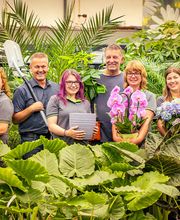 Blumen-Risse Team aus der Abteilung Floristik, Blumenmarkt Lüdinghausen, Baumschule, Keramik, Zimmerpflanzen und Beetware steht zwischen grünen Zimmerpflanzen im Blumenmarkt Lüdinghausen.