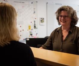 Eine Frau mit lockigem Haar und Brille steht lächelnd hinter einem Empfangstresen. Ihr gegenüber ist der Rücken einer anderen Person zu sehen. Im Hintergrund hängen ein Jahresplaner und eine Pinnwand an der Wand. Der Raum wirkt hell und freundlich.