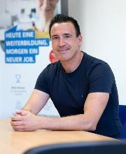 Ein Mann mit kurzen, schwarzen Haaren und dunkelblauem T-Shirt sitzt an einem Tisch.