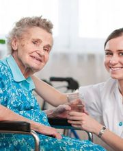 Liebevolle Unterstützung im Alltag: Eine Betreuungskraft begleitet eine Seniorin im Rollstuhl und reicht ihr ein Glas Wasser. DOPAS Pflege steht für persönliche Seniorenbetreuung und Unterstützung im eigenen Zuhause in Münster und Umgebung.