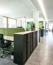 Ein Schulungsraum mit mehreren Arbeitsbereichen, die durch grüne Trennwände voneinander getrennt sind. Jeder Arbeitsplatz besteht aus 2 PC-Bildschirmen, Tastatur und Maus sowie Headset. Im Hintergrund Fototapete von einem Wald. Schwarze Büroschränke.