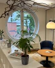 Ein gemütlicher Aufenthaltsraum mit schwarzen Ledersesseln, einem runden Tisch, einem weißen Regal auf dem grüne Zimmerpflanzen stehen. Der Raum hat ein großes, rundes Fenster und es hängt ein Bild von einem Leuchtturm an der Wand.