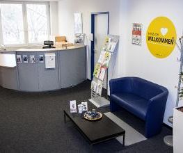 Eingangsbereich. Es zeigt eine Sitzgelegenheit, bestehend aus einem blauen Ledersofa und einem schwarzen Beistelltisch, und den Empfangsthresen..