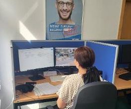Frau sitzt an einem Arbeitsplatz mit 2 Bildschirmen, Headset und Tastatur. Vor ihr liegen mehrere Zettel, an der Wand über ihr hängt ein Plakat.