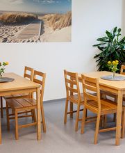 Heller Pausenraum mit hellbraunen Holztischen auf denen gelbe Blumen stehen, Stühlen und einer grünen Zimmerpflanze. An der Wand hängt ein Bild von einem Strand.