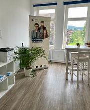 ein heller Aufenthaltsraum mir 2 großen Fenstern. Rechts steht ein weißer Tisch mit Bar-Hockern. Links steht ein Regal, auf dem ein Drucker steht. Daneben steht eine Zimmerpflanze und ein Roll Up der WBS GRUPPE