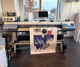 mediaService Bremen | Full-Stack Werbeagentur - Ein professioneller Roland VersaCAMM Plotter und Großformatdrucker druckt ein großes Ausstellungsplakat in einer hellen Druckwerkstatt.