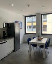 Eine Küche mit Mikrowelle, Kaffeemaschine, Kühlschrank und einem weißen Tisch mit 4 Stühlen, auf dem eine Blume und eine Obstschale steht.