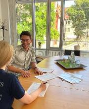 2 Personen sitzen an einem langen Tisch. Vor ihnen mehrere Dokumente. Im Hintergrund kann man durch große Fenster auf grüne Bäume blicken.