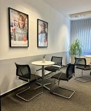 Pausenraum mit Tischen und Stühlen. An der Wand hängen 2 Plakate und im Hintergrund steht eine Pflanze.