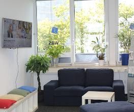 Wartebereich mit Blick auf grüne Bäume. Ein blaues Sofa steht vor dem Fenster mit Pflanzen auf dem Fensterbrett. Rechts steht ein Flyerständer, links hängt ein TV, darunter eine Sitzbank mit bunten Kissen.