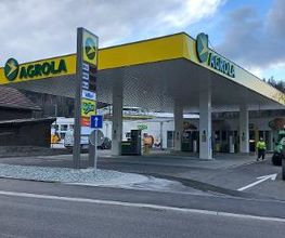 AGROLA Tankstelle in Zell, Luzern, Einfahrt im Vordergrund mit Pfeilmarkierung, links Totem mit AGROLA Diskus Logo, digitaler Preisanzeige, AdBlue, TopShop Logo darunter; im Hintergrund TopShop