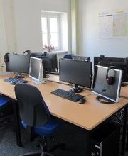 Schulungsraum mit 4 Arbeitsplätzen mit jeweils 2 Bildschirmen, einem Bürostuhl, Tastatur, Maus und Headset. An der Wand hängt ein Wandkalender.