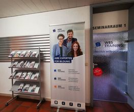 Flur mit Glastür zum Seminarraum. Links steht ein Flyerständer und ein Roll Up Banner der WBS GRUPPE. Rechts die Glastür mit Aufkleber "Herzlich Willkommen"