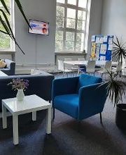 Aufenthaltsraum mit blauen Sesseln, grauen Sofas, einem Tisch mit 7 Stühlen, Fernseher an der Wand sowie großen Fenstern, Zimmerpflanzen und einer Moderationstafel.