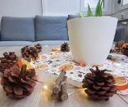 dekorierter Beistelltisch mit Lichterkette, Tannenzapfen und einer herbstlichen Servierte unter einem Blumentopf. Im Hintergrund steht ein graues Sofa.