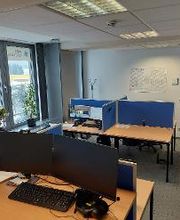 Raum mit mehreren Arbeitsplätzen mit jeweils 2 Bildschirmen, einem Bürostuhl, Tastatur und Maus. Links eine Fensterfront mit Zimmerpflanzen, im Hintergrund ein Kalender an der Wand. Der Boden ist mit blauem Teppich ausgelegt.