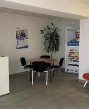 Eingangsbereich mit einer Theke, einem Tisch mit 4 schwarzen Stühlen und 2 Roll up Banner. An der Wand ein Plakat und rechts ein roter Stuhl.