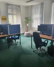 Schulungsraum mit mehreren Arbeitsplätzen. Jeweils mit 2 Bildschirmen, einem Sessel, Tastatur und Maus. In der Ecke steht eine Pflanze hinter einem Sitzsack.