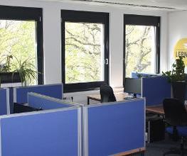 Lernplätze die durch blaue Trennwände voneinander getrennt sind. Jeder Lernplatz besteht aus 2 PC-Bildschirmen, Tastatur und Maus sowie Headset. Fensterfront mit Blick ins Grüne.