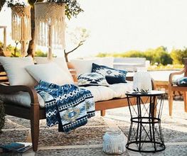 Eine stilvoll eingerichtete Terrasse mit Holzmöbeln, Textilien in Blau-Weiß und mediterranen Details lädt zum Entspannen ein. Das warme Sonnenlicht und die natürliche Umgebung erzeugen ein Gefühl von Urlaub und Leichtigkeit.