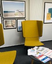 Empfangsbereich im Standort mit zwei  gelben Sesseln und einem Beistelltisch, worauf Zeitschriften liegen. Im Hintergrund sind mehrere Bilder an der Wand zu sehen.