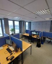 Raum mit mehreren Arbeitsplätzen mit jeweils 2 Bildschirmen, einem Bürostuhl, Tastatur und Maus. Im Hintergrund sind mehrere Schränke zu sehen, links eine Fensterfront mit Zimmerpflanzen. Der Boden ist mit Teppich ausgelegt.