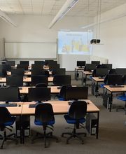 Schulungsraum. Tischreihen mit Bildschirmen und blauen Stühlen. Die Wände sind rot und blau gestrichen. In dem Raum befindet sich ein Whiteboard und an der Wand ist eine Projektion vom Beamer zu sehen.