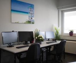 Zwei Arbeitsplätze für Teilnehmedne mit 2 Bildschrimen, Tastatur, Maus und Headset. An der Wand hängt ein schönes Strandbild und auf den Tischen stehen Blumen.