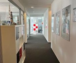 Ein heller Flur mit dunklem Teppichboden. Rechts hängen Poster an der weißen Wand, links stehen Regale mit Fächern. Am Ende des Flurs ist eine Glastür mit einer rot-weißen Markierung und ein Fluchtwegschild zu sehen.