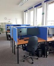 Arbeitsplätze mit jeweils einem Bürostuhl, 2 Bildschirmen und Tastatur und Maus. Im Hintergrund ein Regal mit Zimmerpflanzen. Rechts mehrere Fenster.