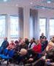 Eine Frau hält einen Vortrag und steht an einem Rednerpult. Vor ihr sitzen ca. 50 bis 80 Personen, die dem Vortrag folgen.