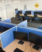 Ein Schulungsraum mit mehreren Arbeitsbereichen, die durch blaue Trennwände voneinander getrennt sind. Jeder Arbeitsplatz besteht aus 2 PC-Bildschirmen, Tastatur und Maus sowie Headset. Blaue Trennwände.