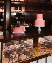 A lit marble and wood display case featuring stacked pink frosted cakes, fresh croissants, delicate tarts, and gourmet tea sandwiches.
