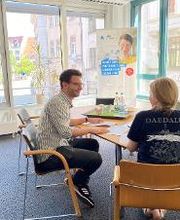 2 Personen sitzen an einem Tisch in einem Lichtdurchfluteten Raum. Im Hintergrund steht ein Roll-Up Banner der WBS TRAINING. Auf dem Tisch ein Tablett mit Wasserflaschen