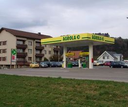 AGROLA Tankstelle in Bütschwil - freistehend mit zwei Tanksäulen. Links Totem mit AGROLA Logo und digitaler Preisanzeige. Dahinter Wohnüberbauung und Parkplatz