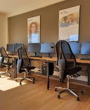 Schulungsraum mit 3 Arbeitsplätzen mit jeweils 2 Bildschirmen, einem Bürostuhl, Tastatur, Maus und Headset. Darüber hängen 2 WBS TRAINING Plakate, daneben eine gelbe Trennwand.