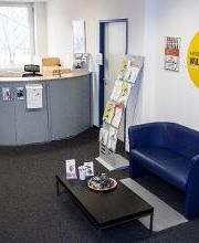 Eingangsbereich. Es zeigt eine Sitzgelegenheit, bestehend aus einem blauen Ledersofa und einem schwarzen Beistelltisch, und den Empfangsthresen..
