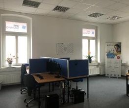 Schulungsraum mit mehreren Arbeitsplätzen mit jeweils 2 Bildschirmen, einem Bürostuhl, Tastatur, Maus und Headset. An der Wand ein Kalender.